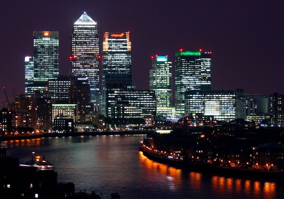 Nighttime view of the Canary Wharf financial district skyline with illuminated modern skyscrapers, including One Canada Square with its distinctive pyramid-shaped top, reflecting on the water of the R