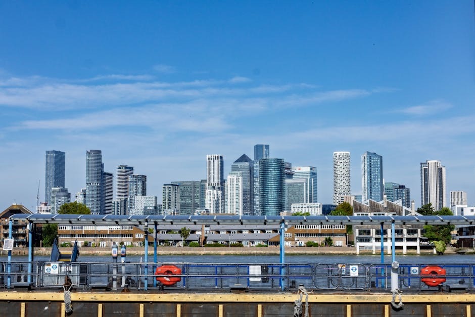The image depicts a modern urban scene with a bright red train crossing a bridge over a calm river, set against a backdrop of tall, contemporary office buildings with glass and steel facades. The trai